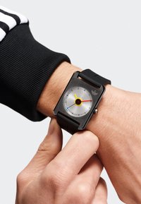 Montre noire avec un cadran métallique rectangulaire, un cadran blanc, des accents jaunes et rouges, un bracelet en silicone noir et un logo visible sur le côté.