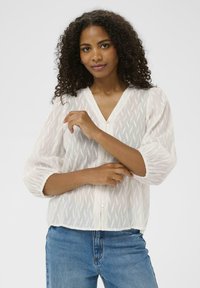 Femme aux cheveux bouclés portant un blouse à col en V texturé blanc et un jean bleu, ajustant sa manche contre un fond clair. 