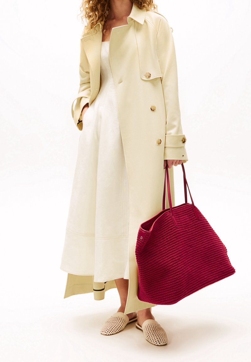Femme portant un trench-coat crème et une robe blanche, tenant un grand sac cabas tressé rouge, avec des chaussures slip-on tressées beige.