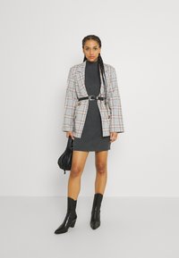 Blazer a cuadros con cinturón, vestido de punto gris ajustado y botines negros. Los accesorios incluyen un pequeño bolso negro. Texturas suaves y formas definidas.
