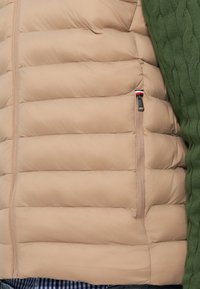 Beige puffer jacket with quilted texture, side zip pocket, and a green knit sleeve. Lightweight and insulated design.