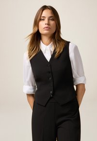 Femeie cu păr șaten deschis, îmbrăcată cu o cămașă albă cu guler sub un vestă neagră cu nasturi și pantaloni negri, pe un fundal simplu.