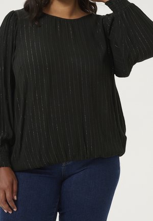 Kvinde iført en sort bluse med lange ærmer med subtile lodrette shimmerstriber og blå jeans, hånd hvilende på hoften og hoved.
