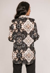 Blouse à manches longues en tissu noir avec un motif floral et géométrique, présentant des accents crème, beige et or, avec une coupe ajustée et un col.