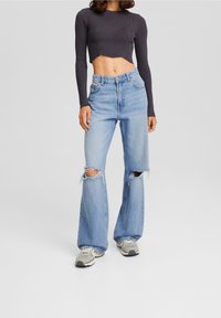 Donna che indossa jeans larghi strappati di colore blu chiaro, un maglione grigio corto con maniche lunghe e sneaker grigie, in piedi contro uno sfondo semplice.