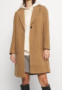 Abrigo de mezcla de lana marrón con solapa con muesca, cierre de un solo botón y dos bolsillos frontales, usado sobre una sudadera crema y una falda estampada.