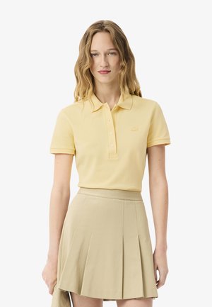 Donna con capelli biondi ondulati che indossa una polo a maniche corte di un giallo pallido e una gonna beige a pieghe, in piedi davanti a uno sfondo semplice.