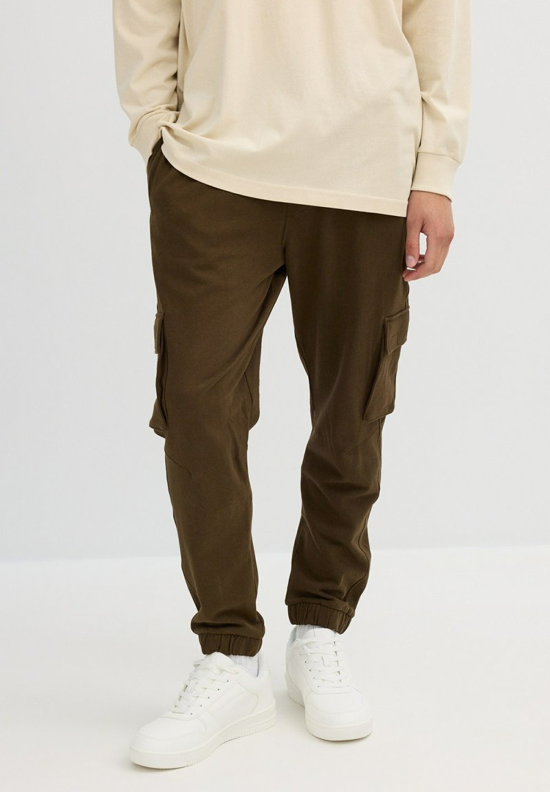 Hombre con pantalones cargo marrón oscuro con bolsillos laterales, camisa de manga larga beige y zapatillas blancas, de pie frente a un fondo liso.
