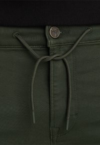 Pantalon vert foncé avec un cordon de serrage noué à la taille et un petit bouton rond en métal au-dessus de la braguette, porté avec un haut noir côtelé.