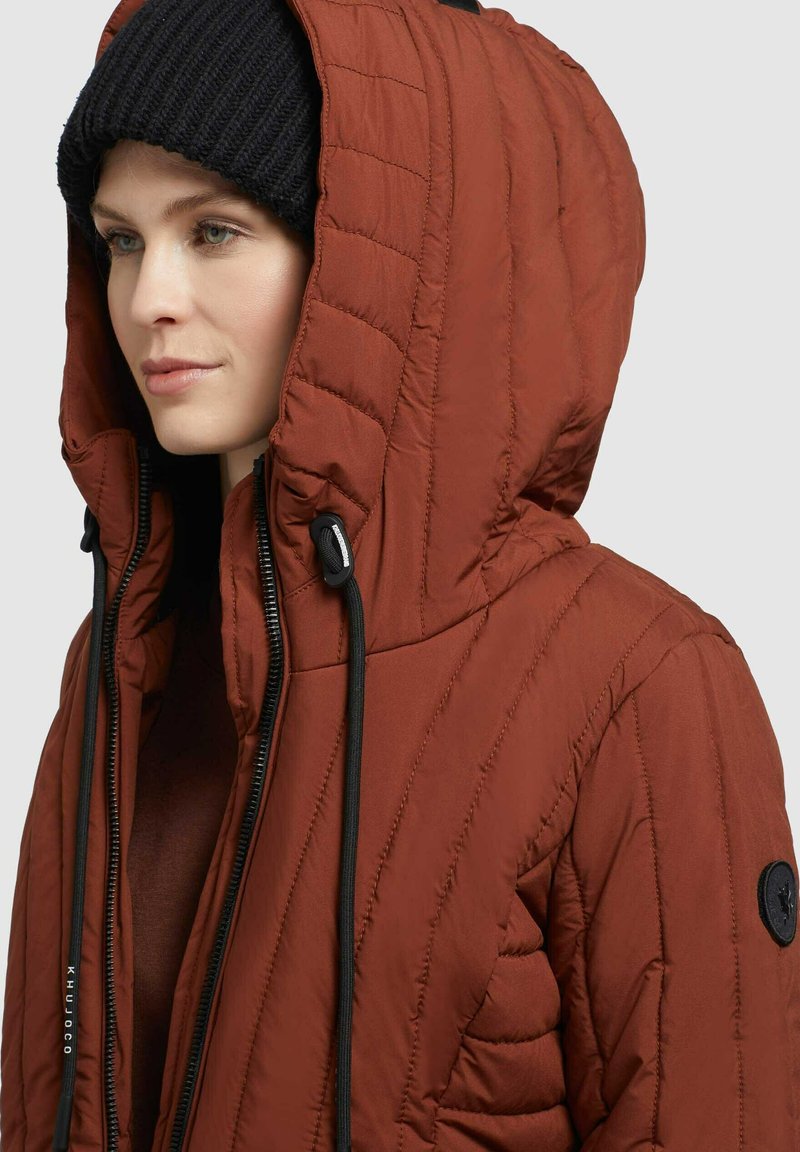 Rote gepolsterte Jacke mit hohem Kragen, ausgestattet mit schwarzen Kordeln und diagonaler Steppung. Eine schwarze gestrickte Mütze sorgt für Kontrast.