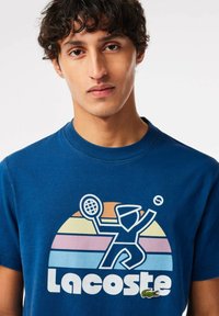 Camiseta de algodón azul con un gráfico retro colorido de tenis, texto "Lacoste" y un pequeño logo de cocodrilo verde en el frente.