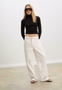 Femme portant un pull noir ample, un pantalon large blanc, des sandales noires et de grandes lunettes de soleil sombres, se tenant sur un sol uni contre un mur blanc.