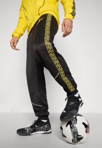 Sorte atletiske bukser med gule mønstrede side striber, parret med sorte sko, hvilende på en fodbold, med en gul hoodie.