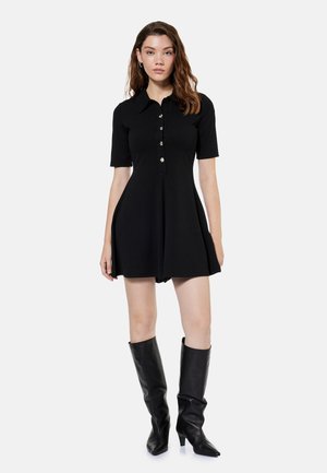 Nainen, joka käyttää lyhyttä mustaa mekkoa, jossa on napit ja kaulus, yhdistettynä polven korkeisiin mustiin nahkasaappaisiin, seisoo yksivärisen taustan edessä.