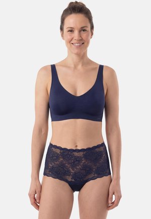 Nainen hymyilee, hänellä on yllään saumaton tummansininen rintaliivit leveillä olkaimilla ja korkeavyötäröiset tummansiniset kukkakirjailulla koristellut pitsialushousut, seisten valkoista taustaa vasten.