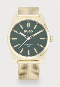 Reloj dorado con esfera verde, que presenta marcadores de hora dorados, manecillas blancas y una pulsera de malla. Resistente al agua hasta 5 bares.