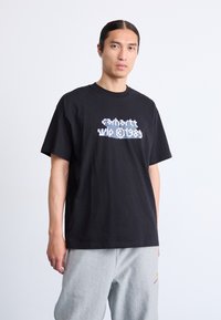 Mand med langt, mørkt hår i en sort grafisk T-shirt og lysegrå sweatpants, der står foran en ensfarvet hvid baggrund.