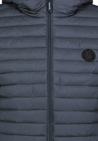 Gestepptes Dunkelgrau-Jackett mit horizontalen Nähten, Reißverschluss und einem runden Logopatch auf der linken Brust. Glattes, leichtes Material.