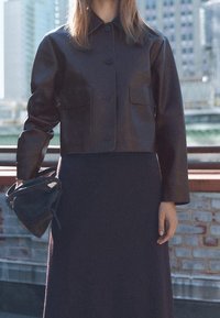 Brązowa skórzana krótka kurtka zapinana na guziki z dwoma przednimi kieszeniami, zestawiona z długą ciemną sukienką i małą czarną torebką.