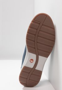 Sula av en blå sko med brun gummisula och vit mellansula, lutande mot en vit vägg och yta.