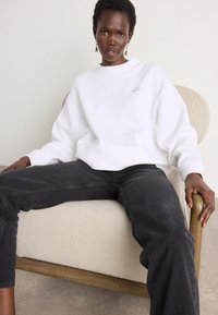 White cotton sweatshirt with a crew neckline and dropped shoulders, featuring a small embroidered logo. Worn with dark gray jeans.