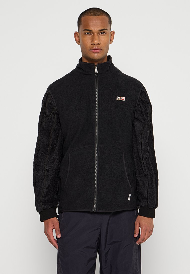 Veste en polaire noire avec fermeture éclair, dotée de manches en tissu texturé noir, d'un col haut et d'un petit logo sur la poitrine.