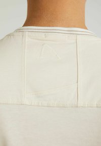 Vaaleanbeige kangas, jossa on teksturoitu kaulus, pieni kolmiomainen ommeltu yksityiskohta ja takana on läppätasku.