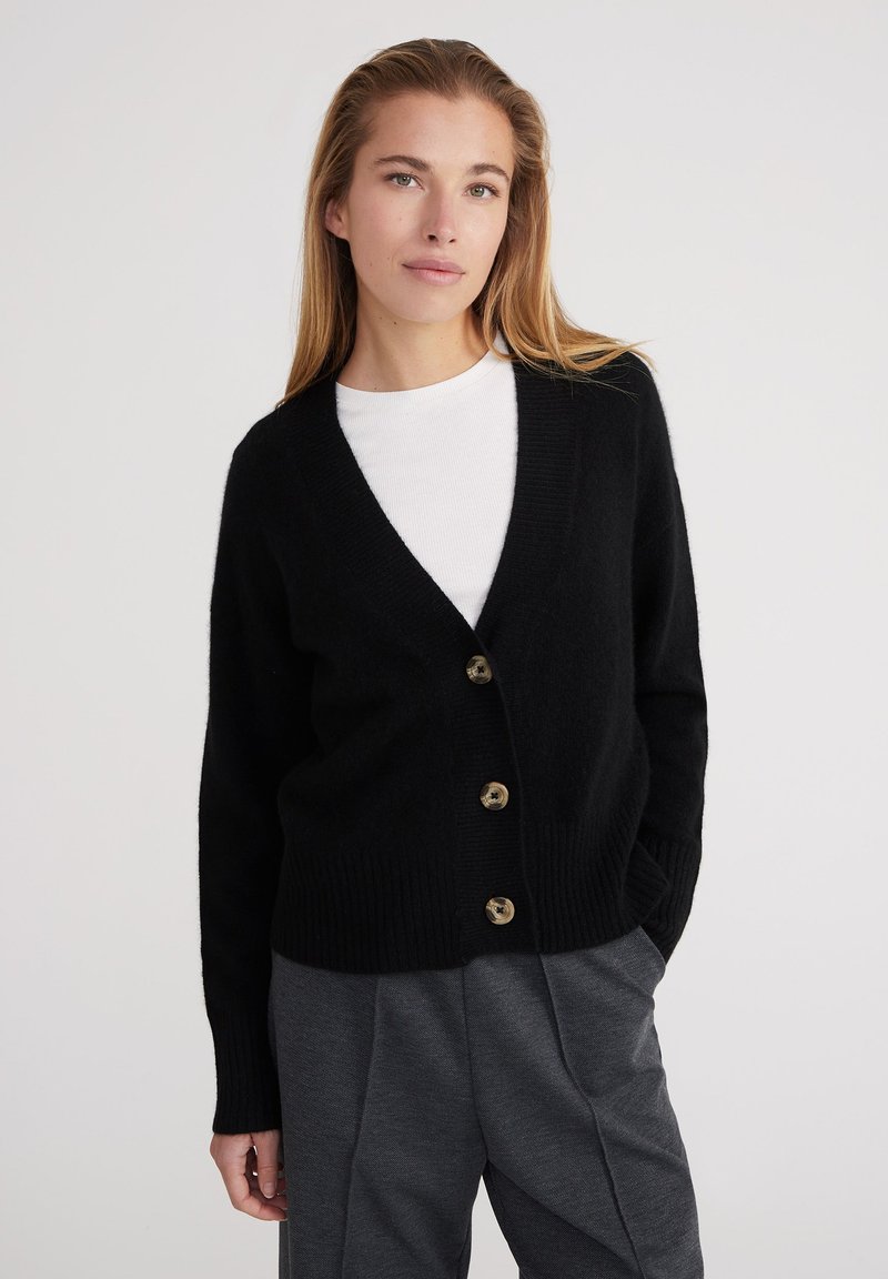 Cárdigan negro con textura acanalada, diseño de escote en V y cinco botones grandes marrones. Lleva sobre una camisa blanca y se combina con pantalones grises.