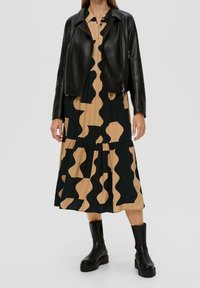 Giubbotto di pelle nera sopra un vestito beige e nero con motivo. Il vestito ha un design a strati e forme ondulate, abbinato a stivali neri con tacco alti fino alla caviglia.