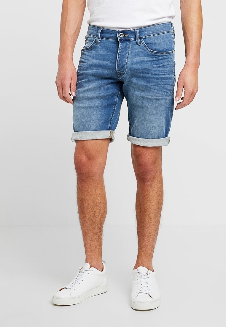 Homme portant un short en denim bleu retroussé, des baskets blanches et un t-shirt blanc, debout devant un fond uni.