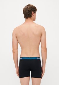 Boxeri negri cu un tiv teal care poartă numele brandului "BOSS" în alb. Materialul pare a fi neted și elastic.