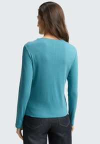 Langsleeve top in effen teal, getailleerde pasvorm, zachte stof, ronde halslijn en subtiele naaddetails op de achterkant. Gecombineerd met donker denim.