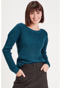 Pull en tricot teal avec un motif texturé, décolleté large et manches longues, associé à un pantalon sombre avec une ceinture.