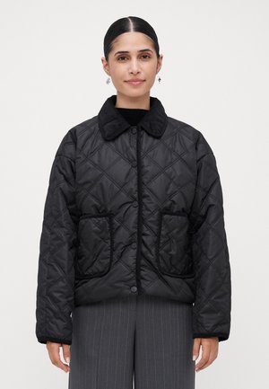 Vrouw met donker haar in een staart, gekleed in een zwarte gewatteerde jas met grote voorzakken en donkere pinstripebroeken, staand tegen een witte achtergrond.