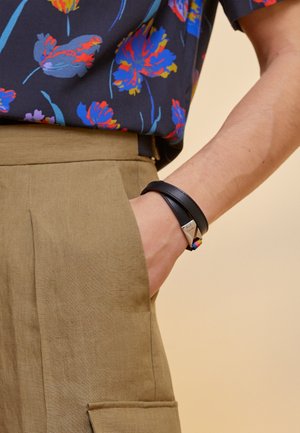 Persoon draagt kaki broek en een zwarte leren wikkelarmband met metalen sluiting, hand nonchalant in de zak, bloemmotief kortärmelig overhemd zichtbaar.