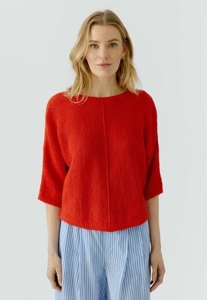 Blonde woman wearing a bright red textured sweater with three-quarter sleeves and light blue and white striped pants, standing against a plain background.