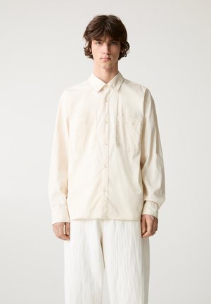 Ljusbeige skjorta med knappar gjord av texturerad tyg, med krage och två bröstfickor, i kombination med vita vida byxor.