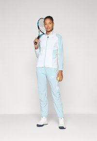 Hellblaues Sport-Jogginganzug mit einer Jacke mit Reißverschluss und passenden Hosen, die weiße Akzente und ein Logo aufweisen. Modell hält einen Tennisschläger und einen Ball.