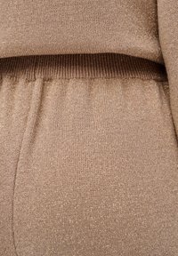 Gros plan d'un tissu tricoté beige avec de fines fibres métalliques, présentant une ceinture côtelée et une texture douce et tombante.
