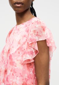 Femme portant un chemisier rose à fleurs avec des manches courtes transparentes et volantées sur un fond clair uni.