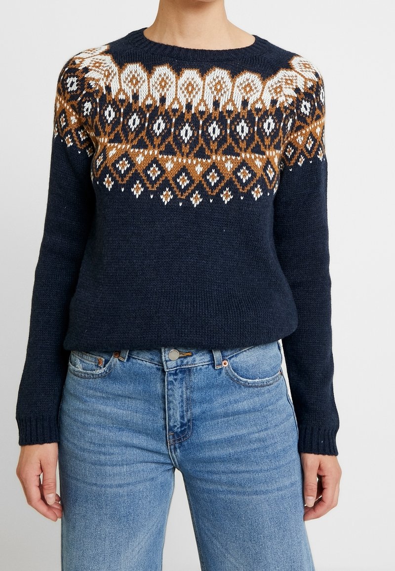Pull en maille bleu marine foncé avec un yoke à motifs comprenant des dessins géométriques en marron, blanc et orange. Poignets et ourlet côtelés.