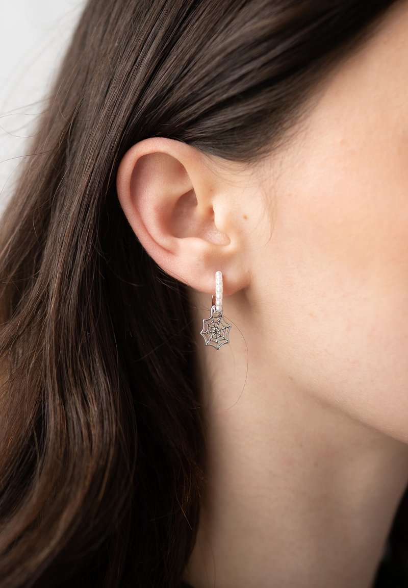 Orecchino a cerchio in argento con superficie texturizzata, caratterizzato da un ciondolo a forma di ragnatela. Piccole perle accentuano il design complessivo.