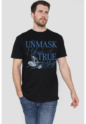 Mann trägt ein schwarzes T-Shirt mit blauem Text "Enthülle dein wahres Ich" sowie kleinen Illustrationen einer Maske und einer Feder, kombiniert mit blauer Jeans.
