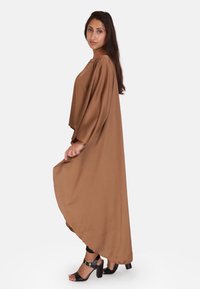 Vestido maxi marrón, fluido, hecho de una tela suave, con mangas largas y un dobladillo redondeado. Combinado con sandalias negras de tacón alto.