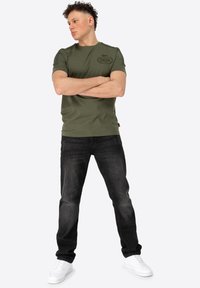 Olivově zelené tričko s krátkým rukávem a texturovaným potiskem loga, spojené s tmavými slim-fit džíny a bílými teniskami. Jednoduchý, neformální vzhled.