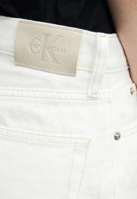 Κοντινό πλάνο λευκών τζιν από τζιν με μπεζ ταμπέλα λογότυπου Calvin Klein στη μέση, φορεμένα με μαύρο τοπ.