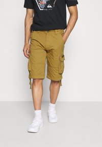 Khaki cargo shorts med flere sidelommer, avslappet passform og oppbrettede kanter. Kombinert med en svart t-skjorte og hvite joggesko.