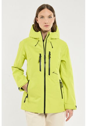 Femme portant une veste à capuche vert citron avec des fermetures éclair noires, debout avec une main dans la poche contre un fond blanc uni.