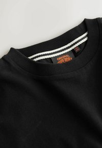 Hanorac negru cu gât rotund și tiv striat, având un accent cu dungi albe. Eticheta afișează „Original Superdry” în portocaliu. Textură moale a materialului.