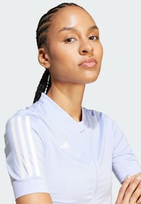 Jasnoniebieska koszulka sportowa z krótkim rękawem, z białymi akcentami w postaci trzech pasków oraz stójką, wykonana z gładkiego, teksturowanego materiału.